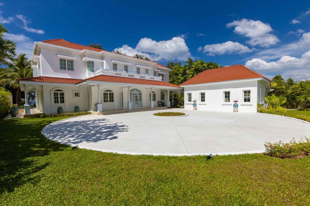 White two-story villa with red-tiled roofs surrounding a circular white driveway and a small landscaped island under a bright blue sky in a tropical setting.