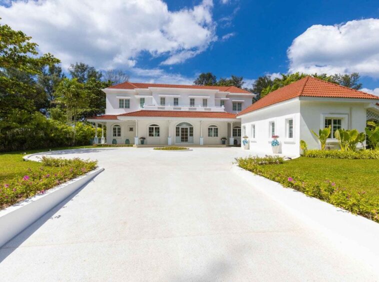 White Mediterranean-style mansion with red-tiled roof and a circular driveway, green lawn and palm trees under a blue sky.