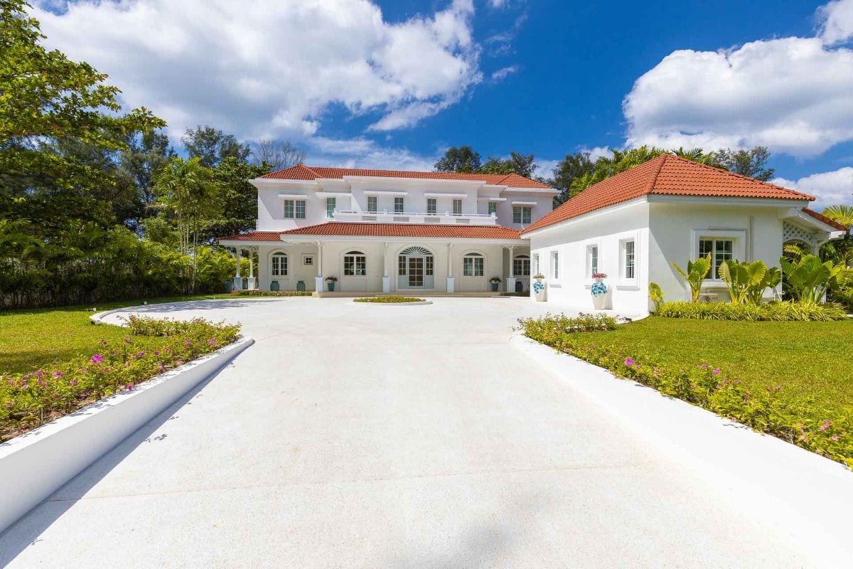 White Mediterranean-style mansion with red-tiled roof and a circular driveway, green lawn and palm trees under a blue sky.
