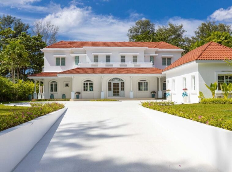 White two-story villa with a red-tiled roof, arched porch, and a manicured tropical landscape under a bright blue sky.
