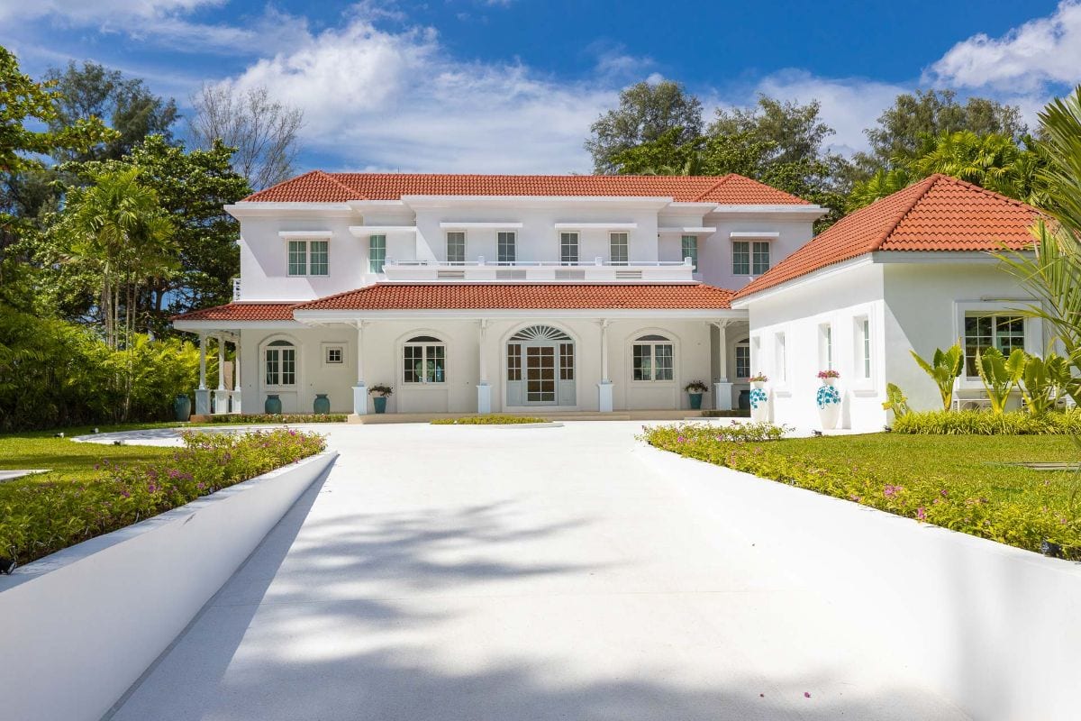White two-story villa with a red-tiled roof, arched porch, and a manicured tropical landscape under a bright blue sky.