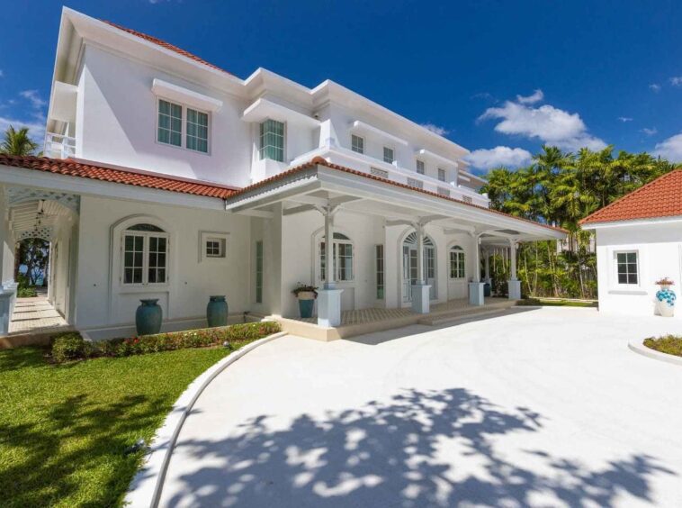 Front view of a white multi-story villa with red tile roof, verandas, and palm trees under a bright blue sky.