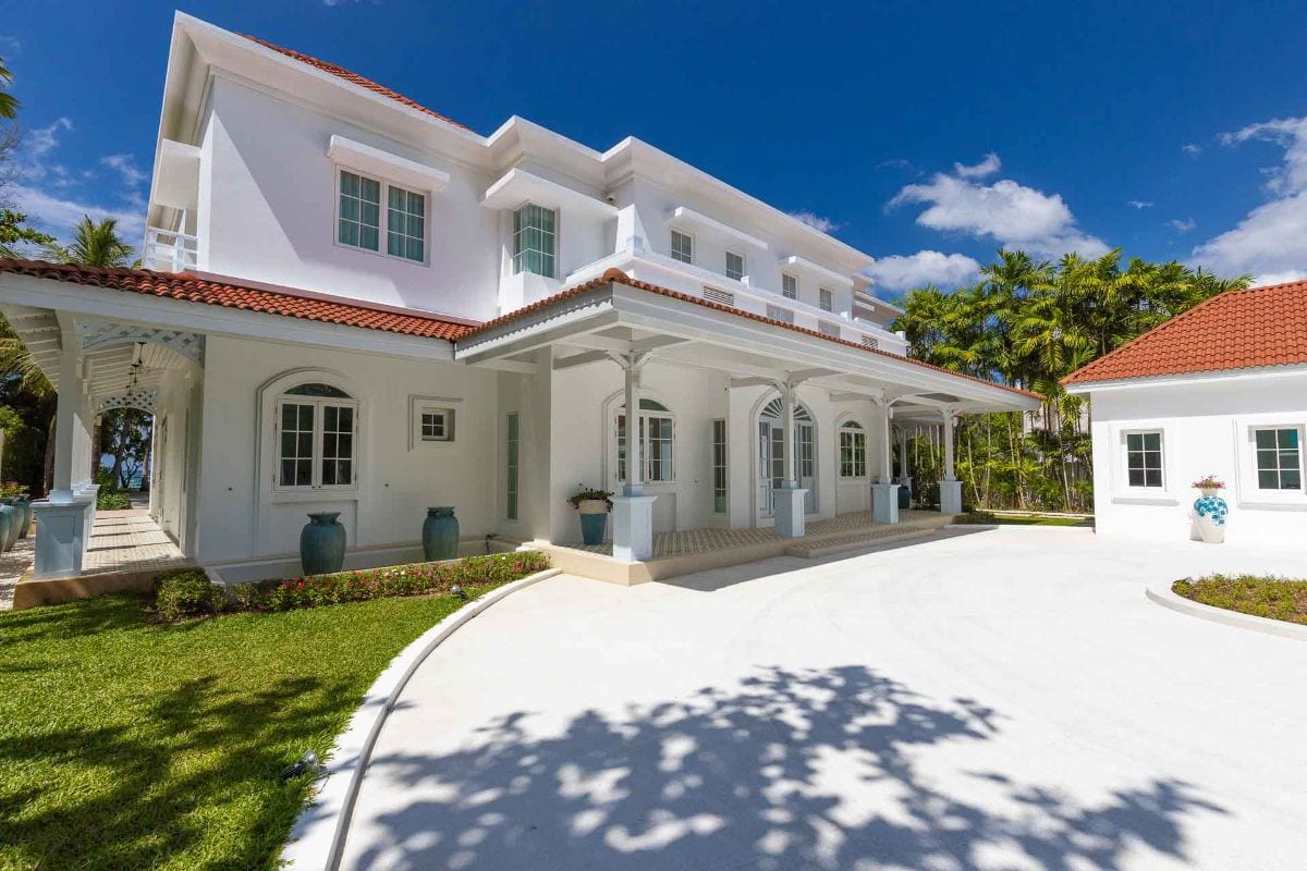 Front view of a white multi-story villa with red tile roof, verandas, and palm trees under a bright blue sky.