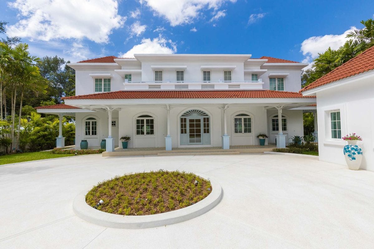 White two-story house with a red-tiled roof, a wide wraparound porch, and pale blue doors, set against a bright blue sky with palm trees on the sides.