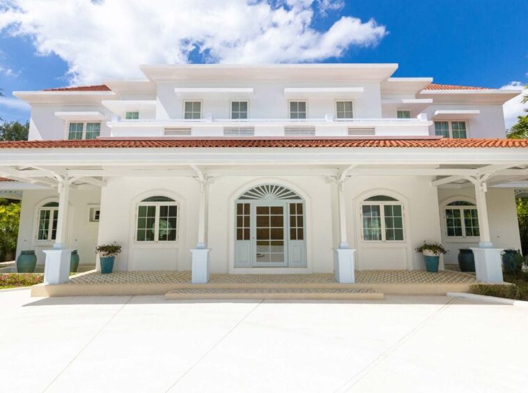 White multi-story house with a red tile roof and large covered front porch under blue sky. A grand white villa with arched entrance, symmetrical windows, and potted plants outside.