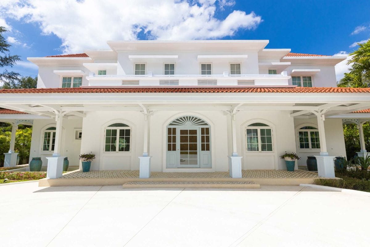 White multi-story house with a red tile roof and large covered front porch under blue sky. A grand white villa with arched entrance, symmetrical windows, and potted plants outside.