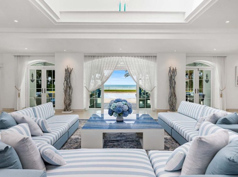 Bright coastal living room with blue-and-white striped sofas arranged around a glass-topped coffee table and a vase of blue hydrangeas, opening to a sea view.
