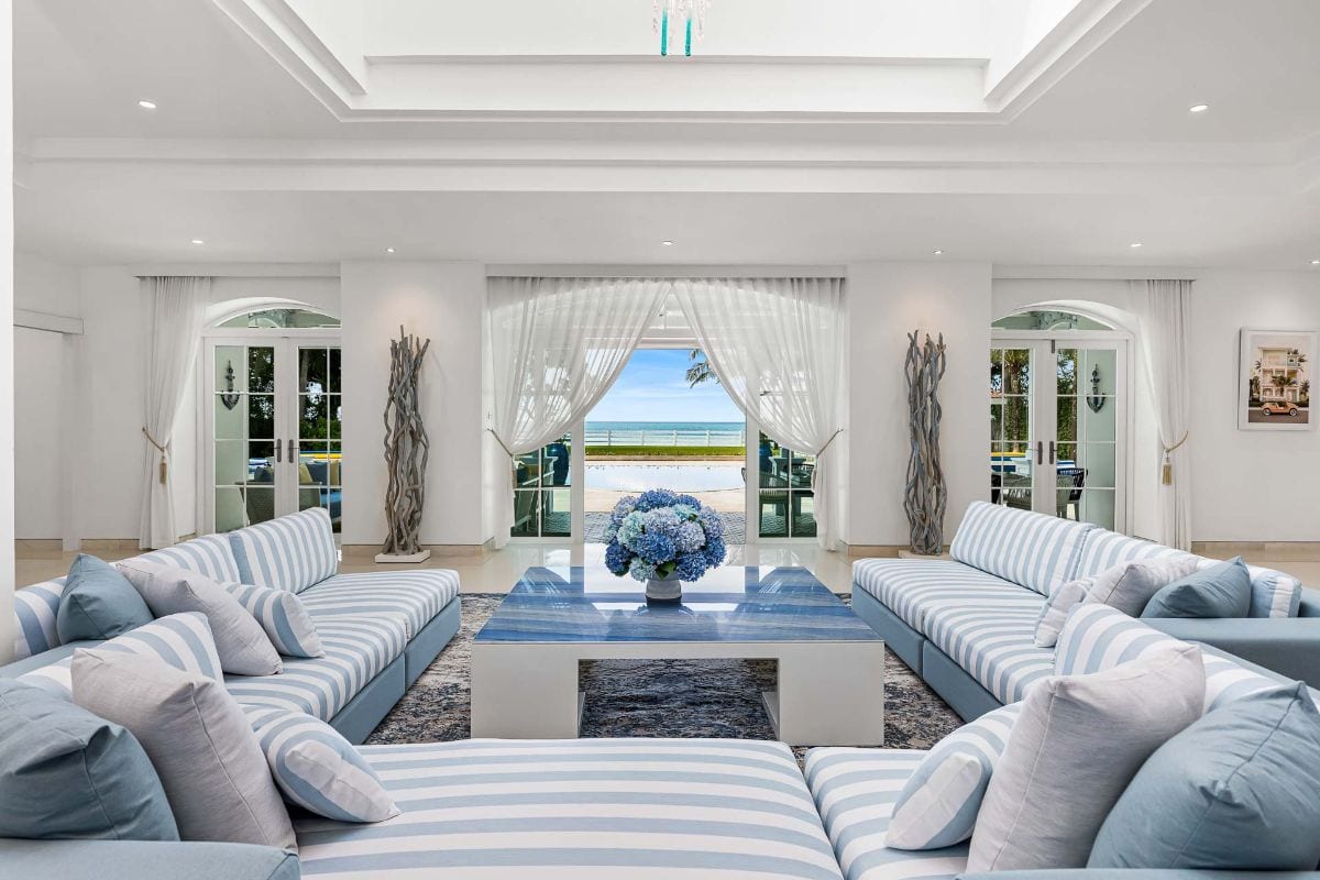 Bright coastal living room with blue-and-white striped sofas arranged around a glass-topped coffee table and a vase of blue hydrangeas, opening to a sea view.