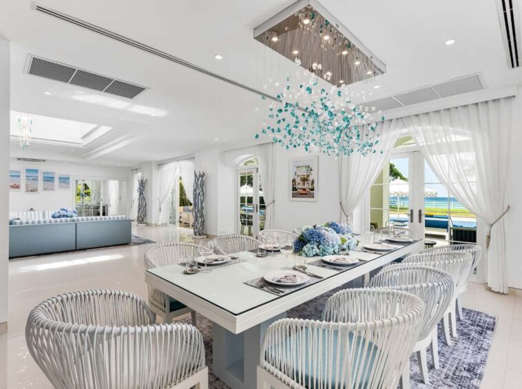 Bright coastal dining room with a long white table, white woven chairs, blue centerpiece, and glass doors opening to an ocean view.