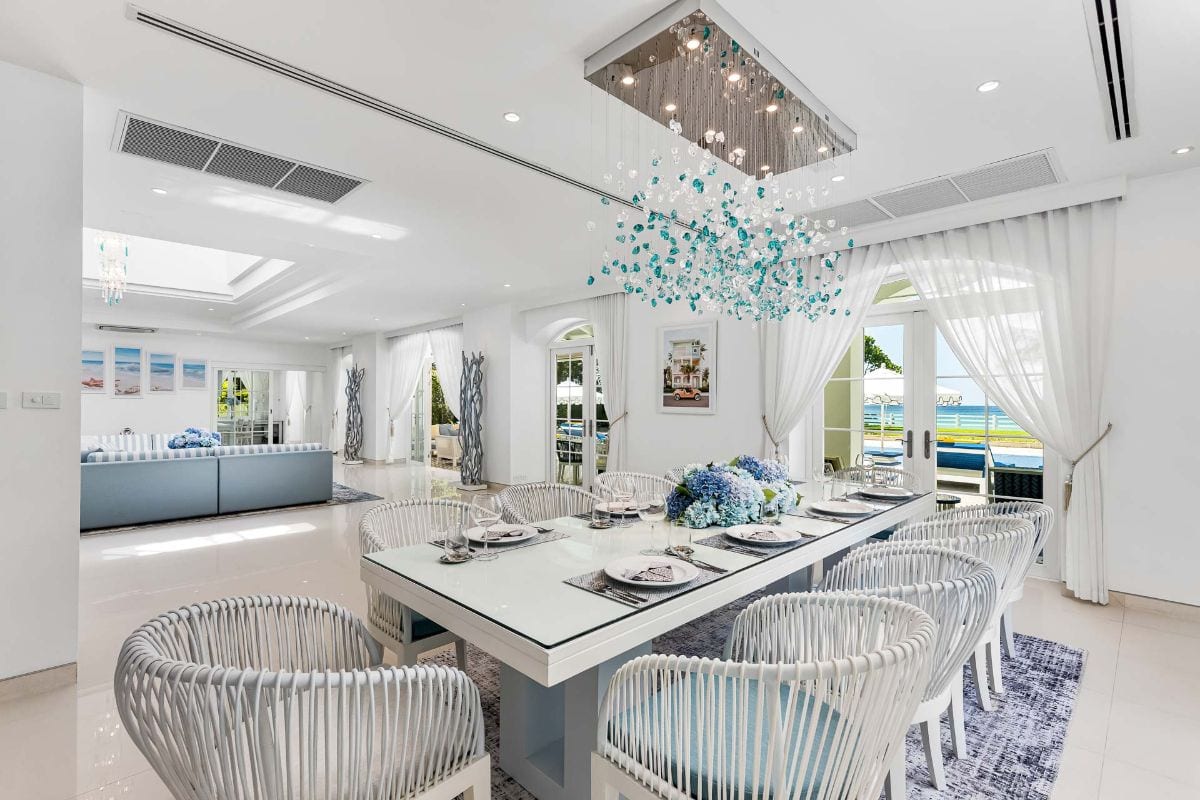 Bright coastal dining room with a long white table, white woven chairs, blue centerpiece, and glass doors opening to an ocean view.