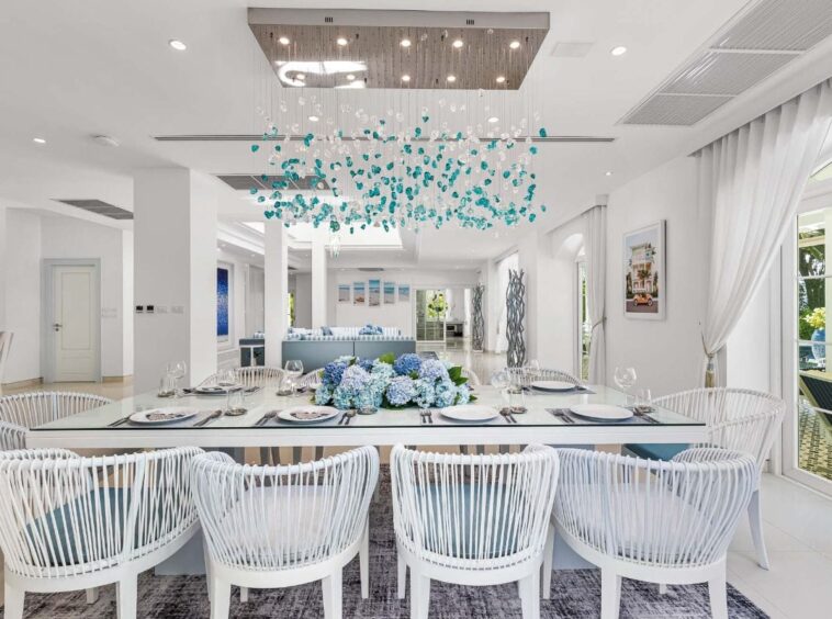 Bright dining room with a long white table, blue hydrangea centerpiece, and white wicker chairs in a sunlit, coastal-style space.