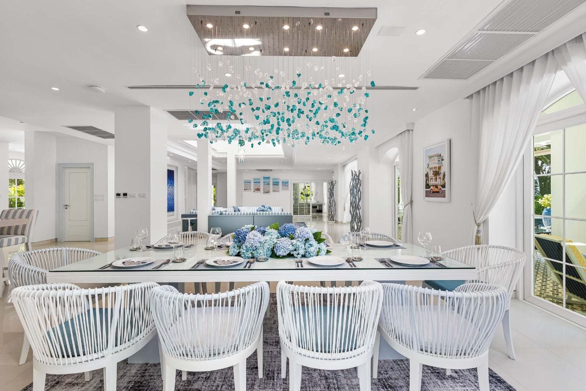 Bright dining room with a long white table, blue hydrangea centerpiece, and white wicker chairs in a sunlit, coastal-style space.