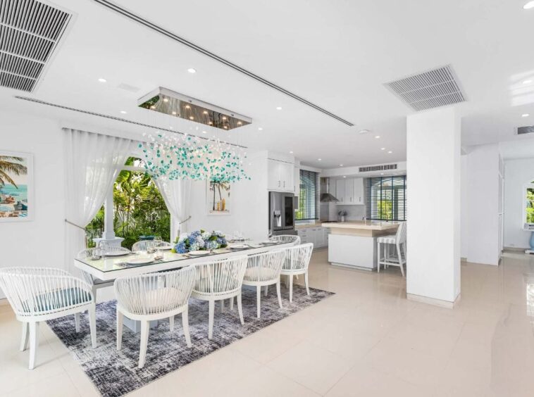 Bright open-plan dining area with a white table and woven chairs, blue floral centerpiece, and a modern kitchen in the background.