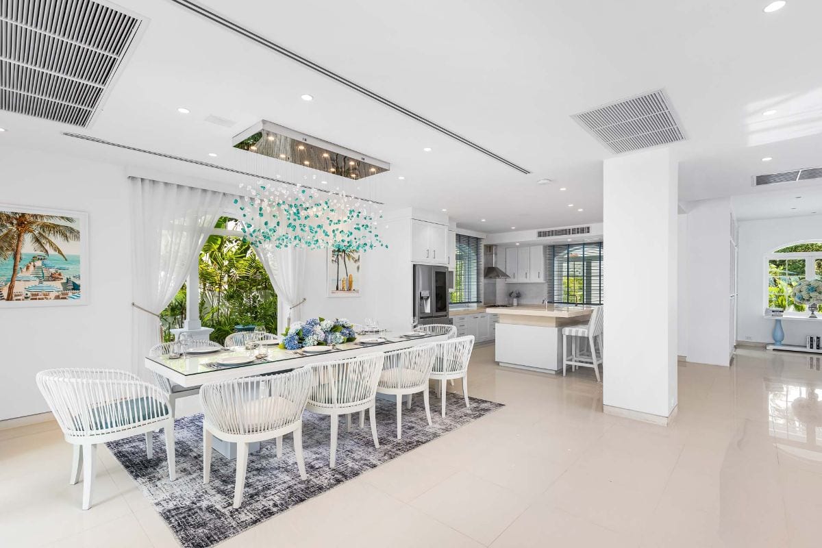 Bright open-plan dining area with a white table and woven chairs, blue floral centerpiece, and a modern kitchen in the background.
