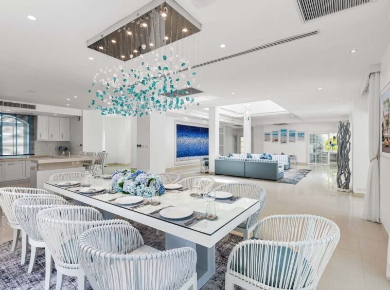 Open-plan dining and living space with a long white table, white woven chairs, and a teal chandelier overhead.