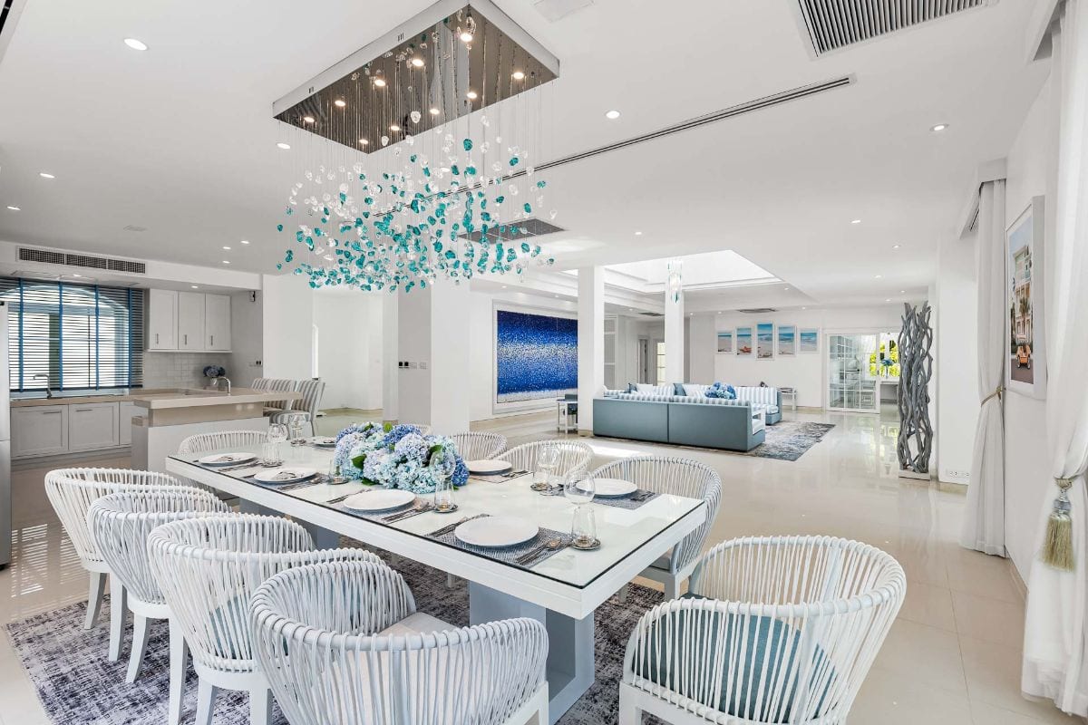 Open-plan dining and living space with a long white table, white woven chairs, and a teal chandelier overhead.