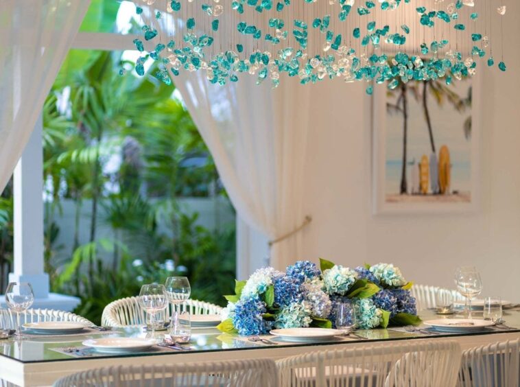 Elegant dining table set with a blue hydrangea centerpiece, glassware, and white plates; white wicker chairs and sheer curtains, with tropical plants outside.