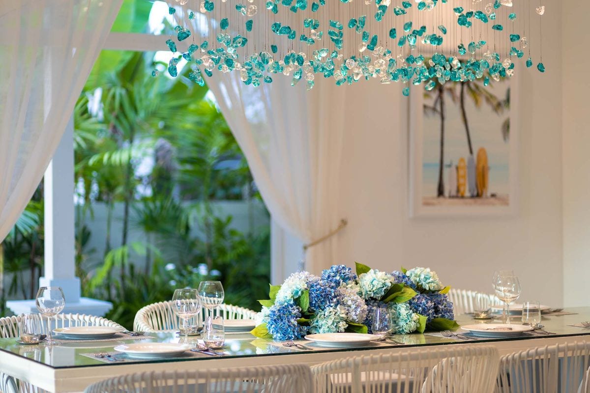 Elegant dining table set with a blue hydrangea centerpiece, glassware, and white plates; white wicker chairs and sheer curtains, with tropical plants outside.