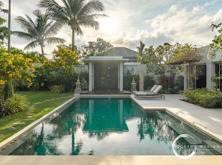 Luxurious backyard with a rectangular pool, white deck, two lounge chairs, and tropical landscaping including palms and yellow flowers. Logo bottom right: Ocean Worldwide.