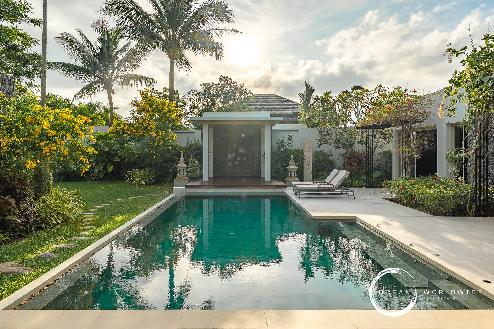 Luxurious backyard with a rectangular pool, white deck, two lounge chairs, and tropical landscaping including palms and yellow flowers. Logo bottom right: Ocean Worldwide.