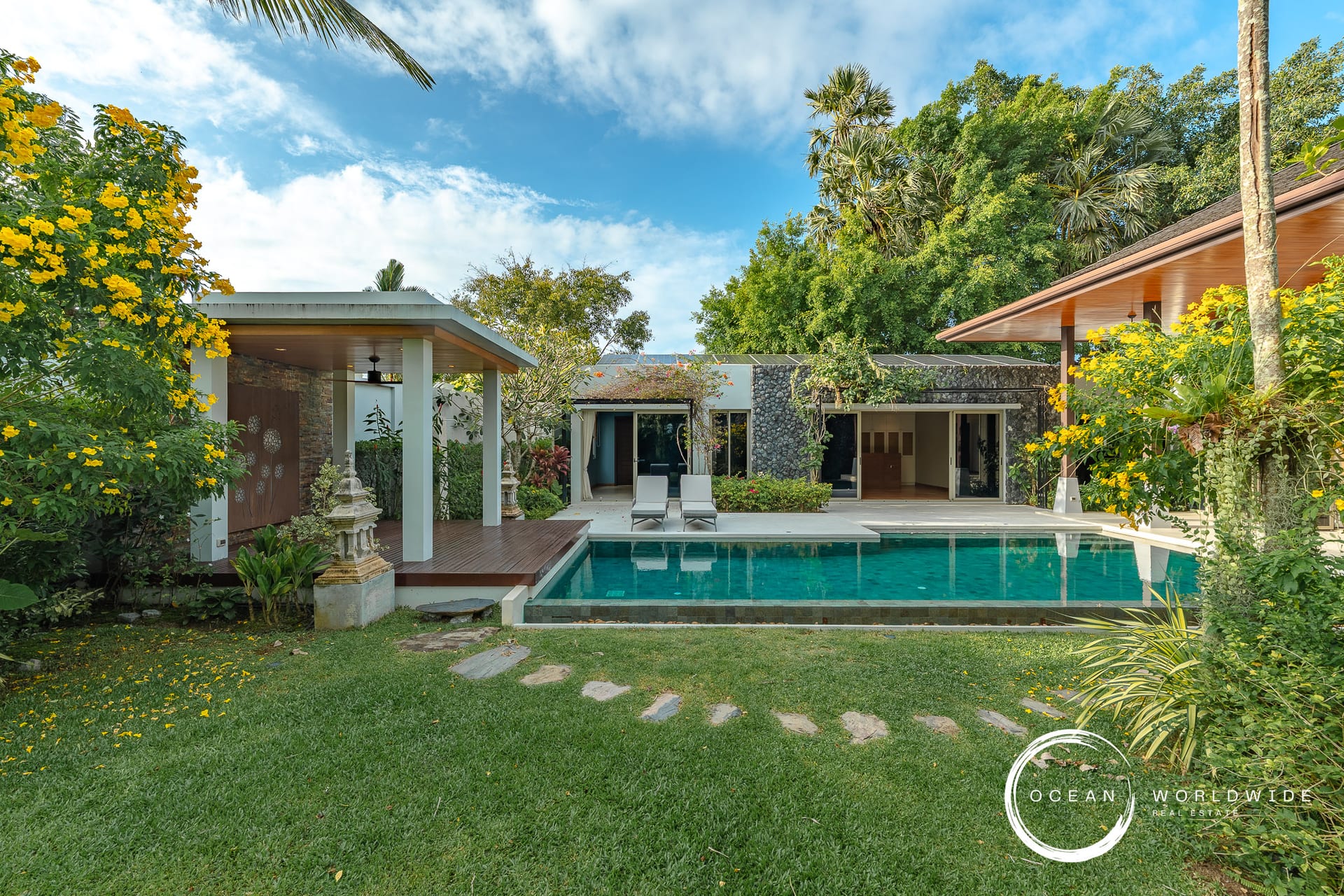 Luxurious villa with a rectangular pool, lounge chairs, and tropical landscaping under a bright blue sky at a resort-style backyard.