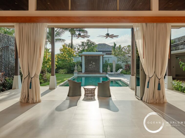 Open-air living area with beige curtains, framing a view of a rectangular pool and lush tropical garden beyond.