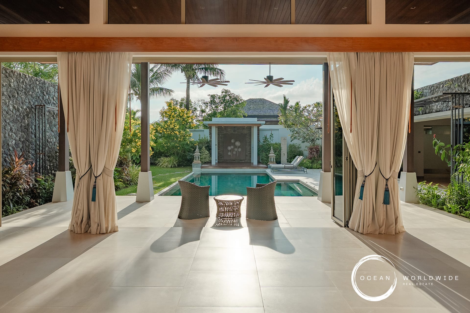 Open-air living area with beige curtains, framing a view of a rectangular pool and lush tropical garden beyond.