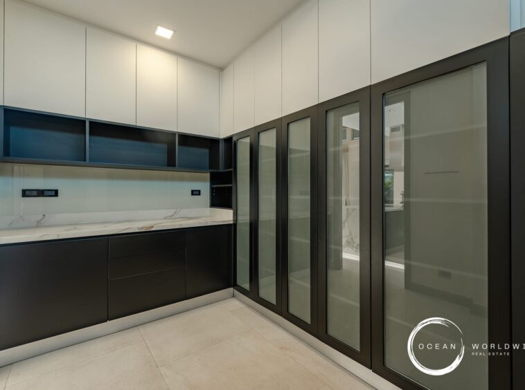 Modern kitchen with white upper cabinets, black base cabinets, marble countertop, blue open shelves, and tall glass-fronted storage along the right wall; watermark logo in bottom-right corner.