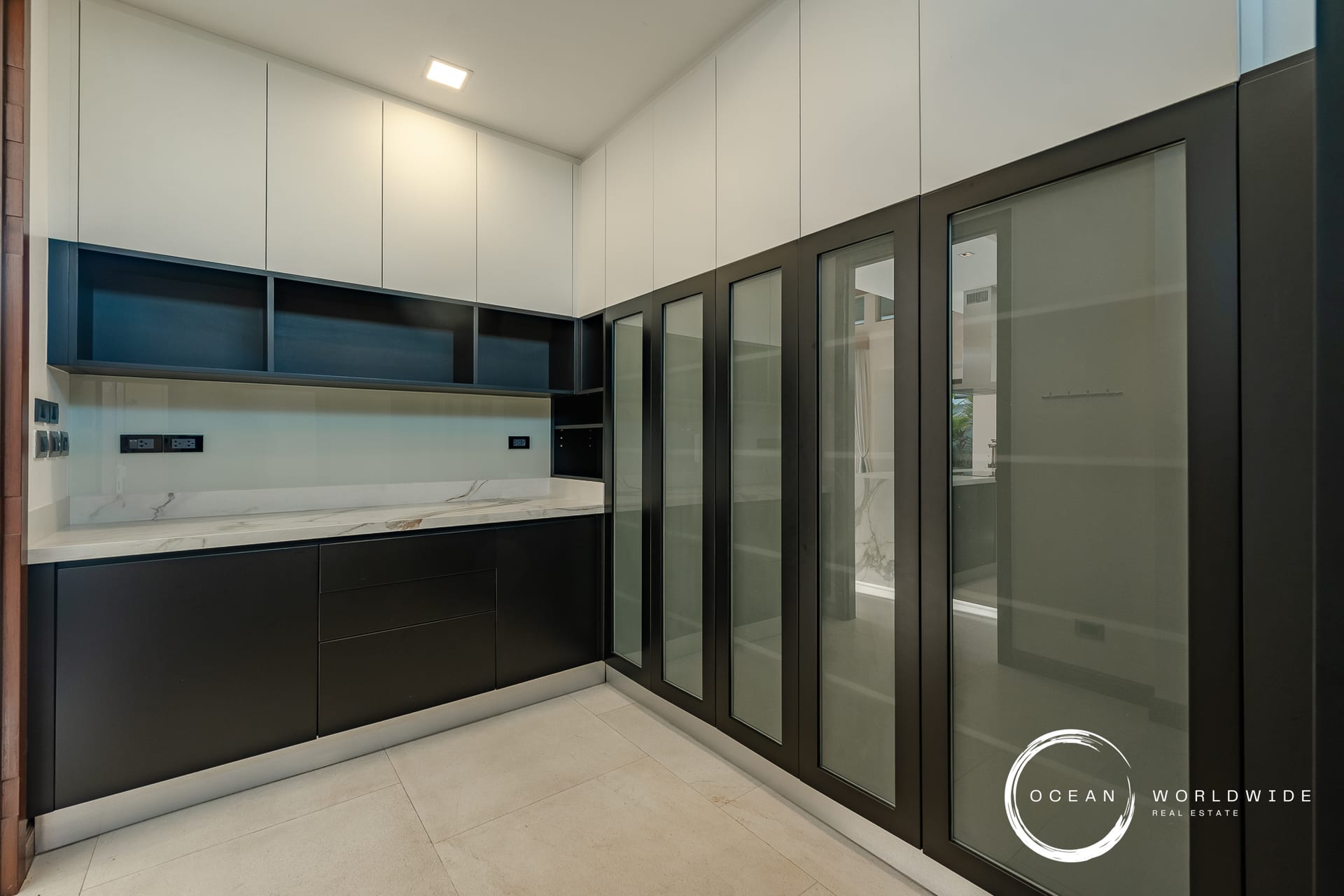 Modern kitchen with white upper cabinets, black base cabinets, marble countertop, blue open shelves, and tall glass-fronted storage along the right wall; watermark logo in bottom-right corner.