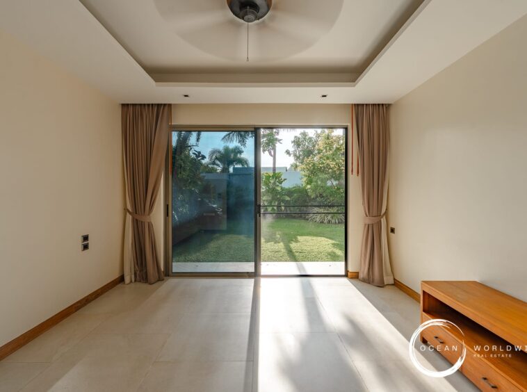 Living room with sliding glass doors to a sunny backyard, beige walls, taupe curtains, and a wooden bench on the right.