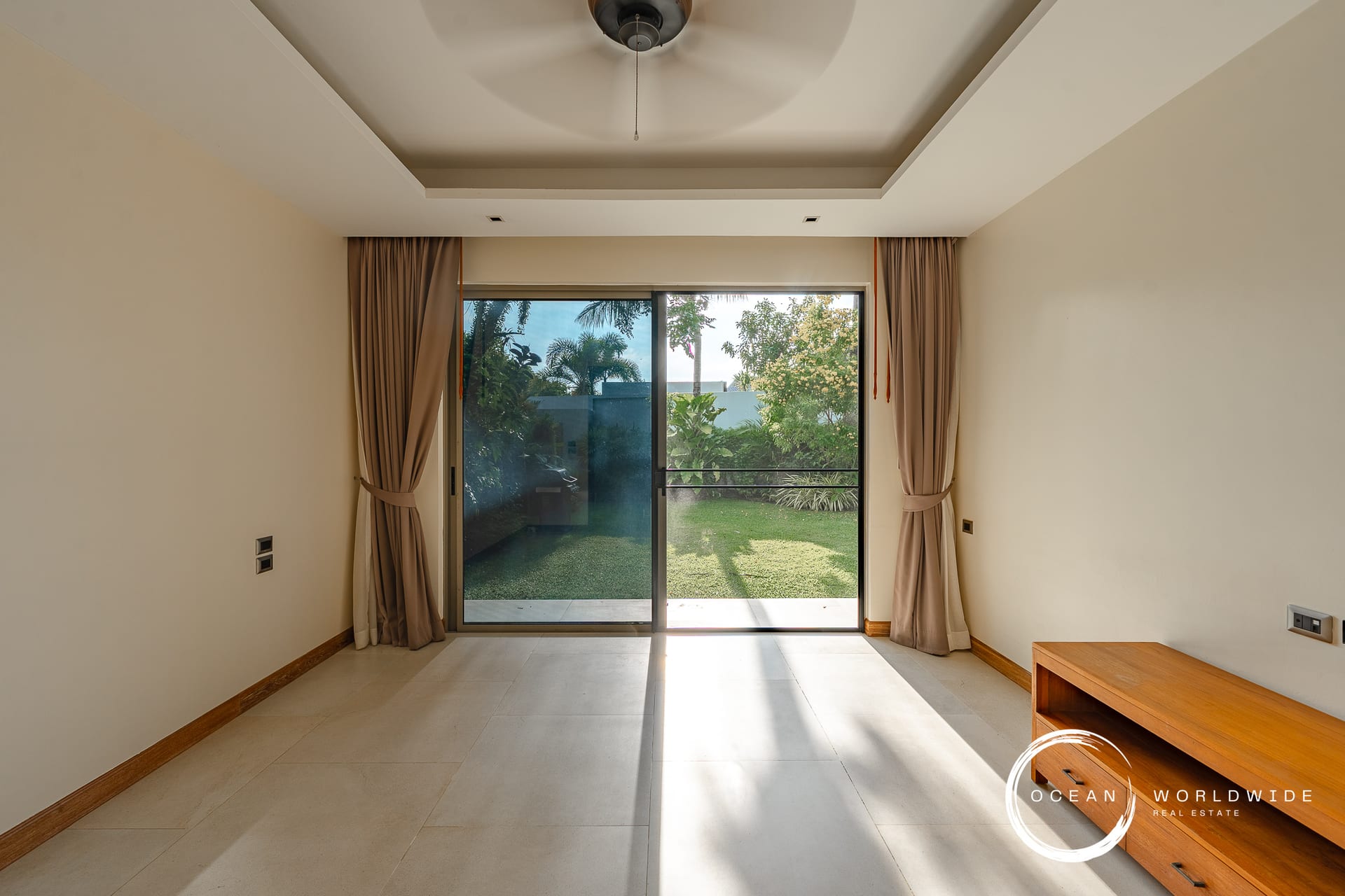Living room with sliding glass doors to a sunny backyard, beige walls, taupe curtains, and a wooden bench on the right.
