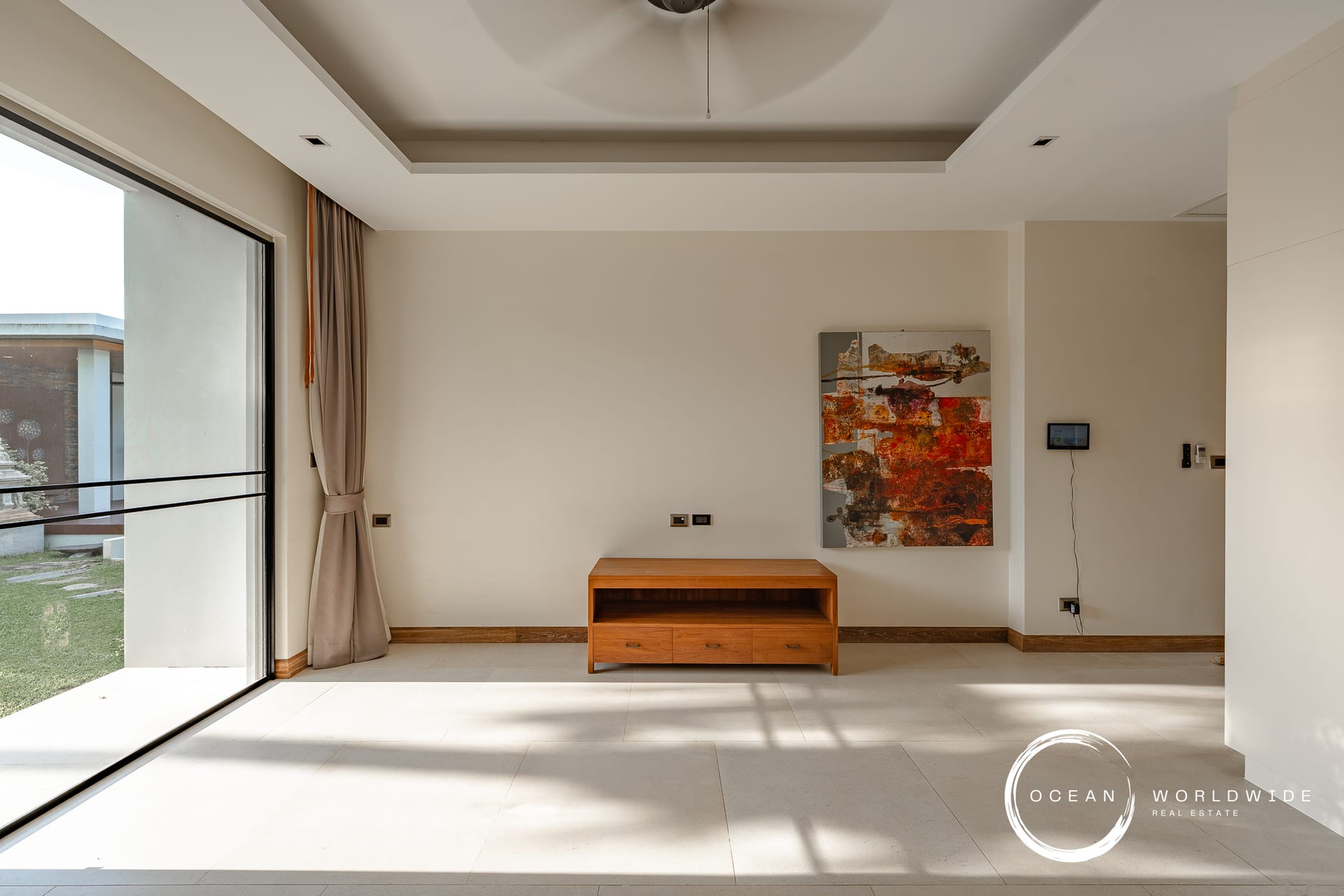 Bright, minimalist living room with a large glass sliding door, beige walls, and a wooden media console beneath an abstract painting.