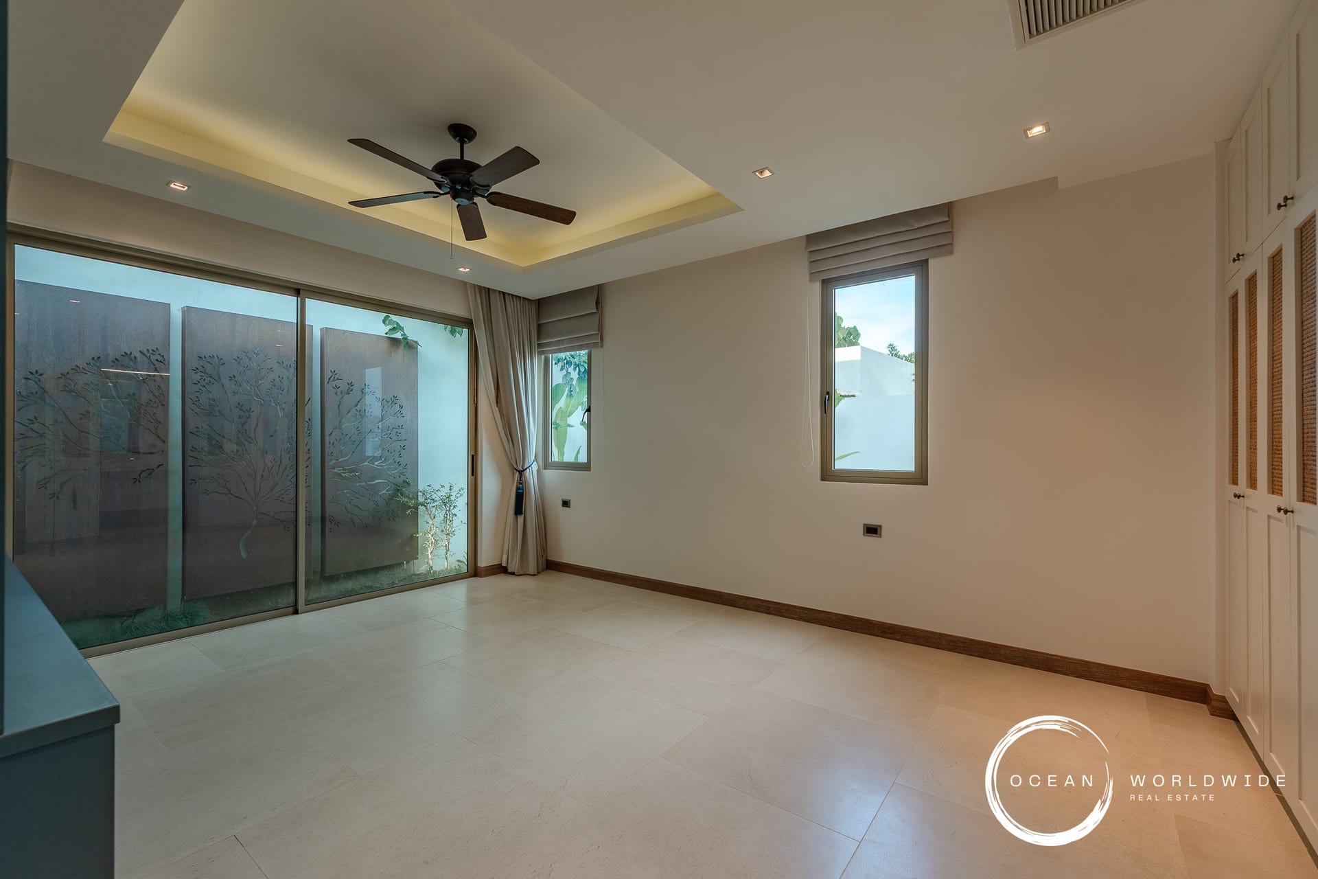 Modern minimalist living room with a ceiling fan, recessed lighting, and large sliding glass doors to an outdoor area. The room is unfurnished with light tile flooring and built-in white cabinets along one wall.