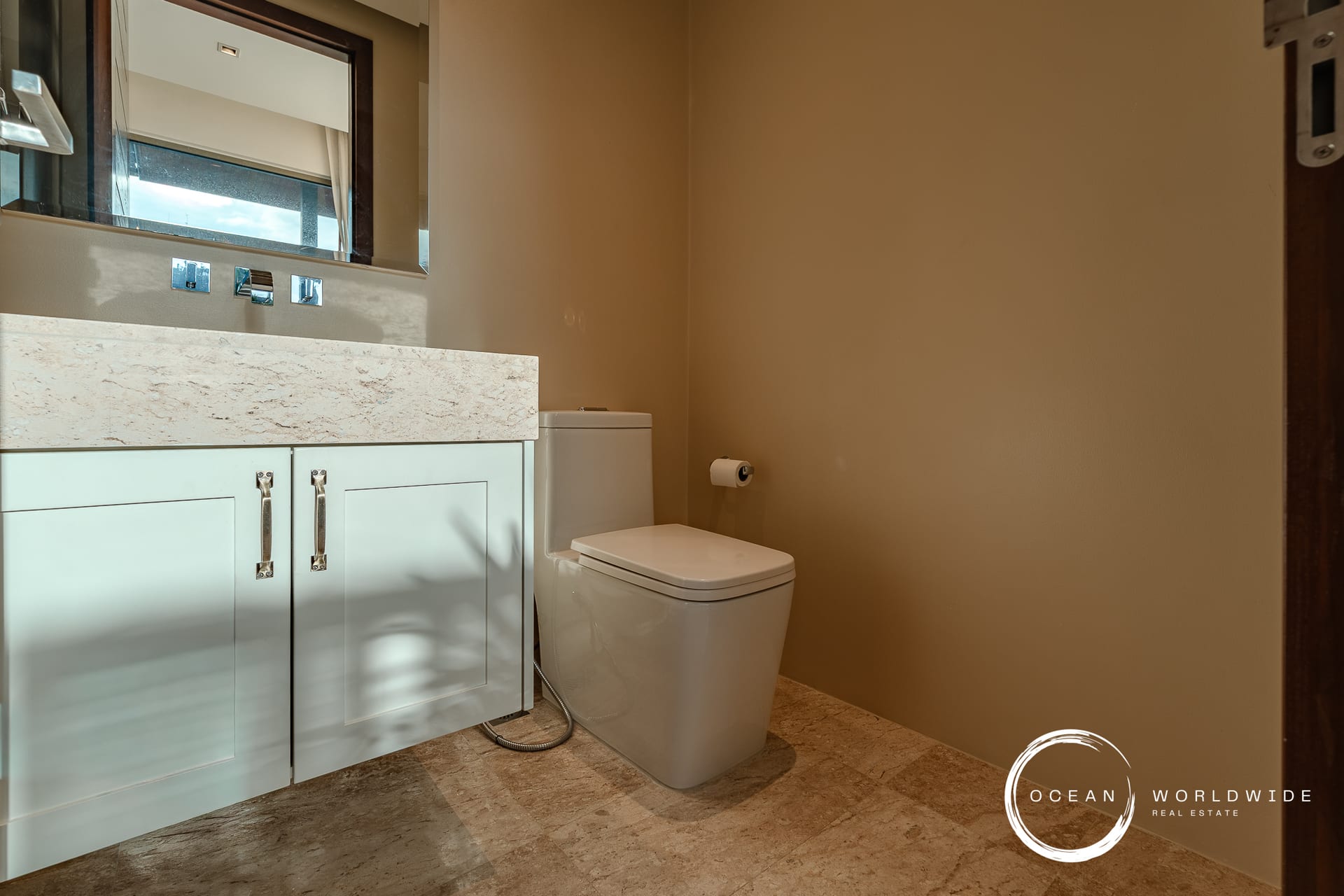 Modern bathroom with a light blue vanity, marble countertop, and a white toilet, glossy chrome fixtures, and a beige wall. Ocean Worldwide logo in corner.