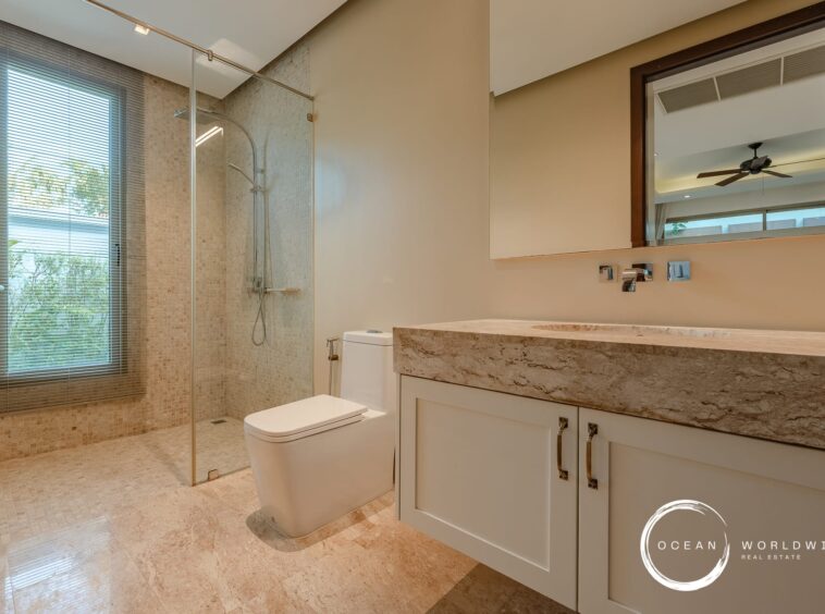 Luxurious beige-tiled bathroom featuring a glass-enclosed shower, large window with blinds, and a marble countertop vanity with white cabinets.