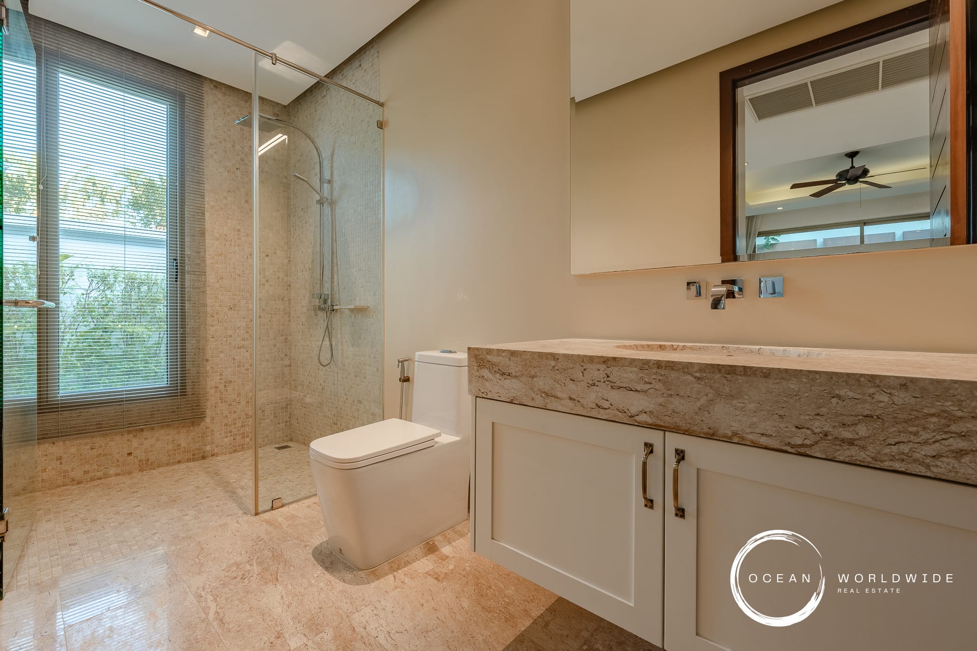 Luxurious beige-tiled bathroom featuring a glass-enclosed shower, large window with blinds, and a marble countertop vanity with white cabinets.