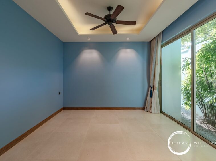Blue-walled empty room with a ceiling fan and large glass sliding doors revealing greenery outside, beige tile floor, and curtain panels on the right.