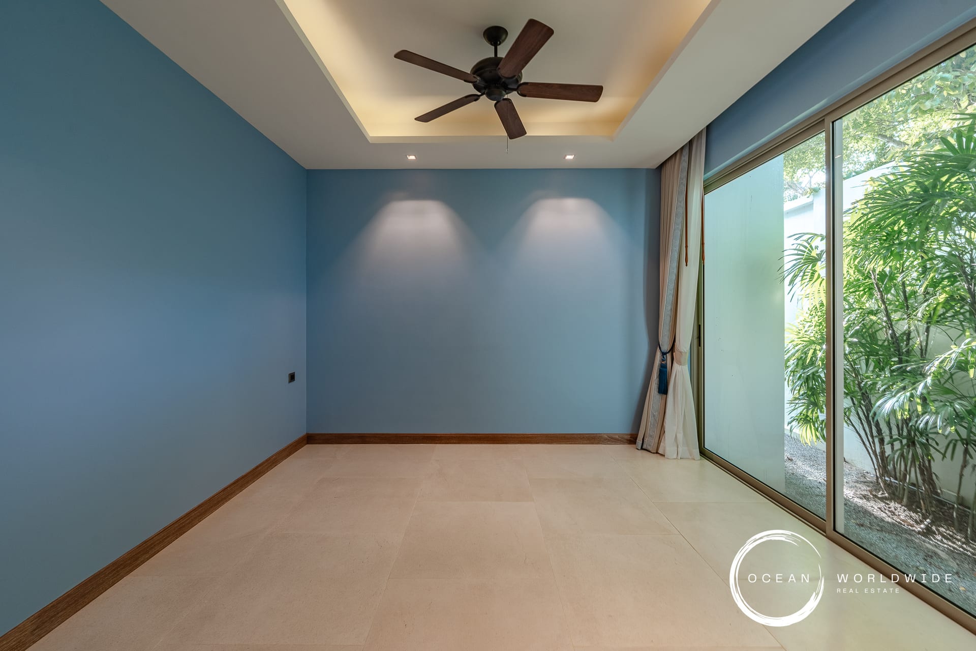 Blue-walled empty room with a ceiling fan and large glass sliding doors revealing greenery outside, beige tile floor, and curtain panels on the right.