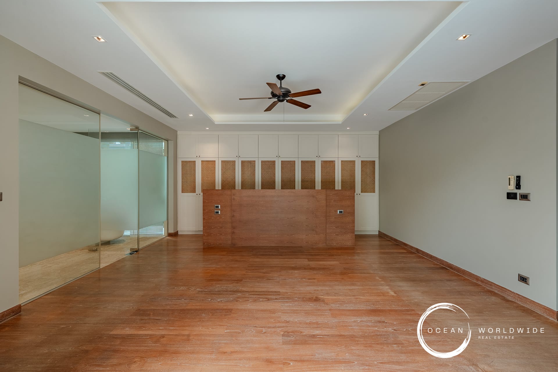 Modern office reception area with white built-in cabinets and a wooden counter, glass partition on the left, and a ceiling fan above.