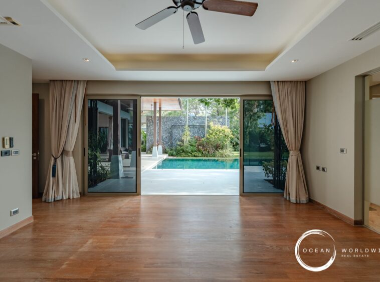 Living area with wood floors and large sliding glass doors opening to a pool and garden, beige walls, and a ceiling fan above.