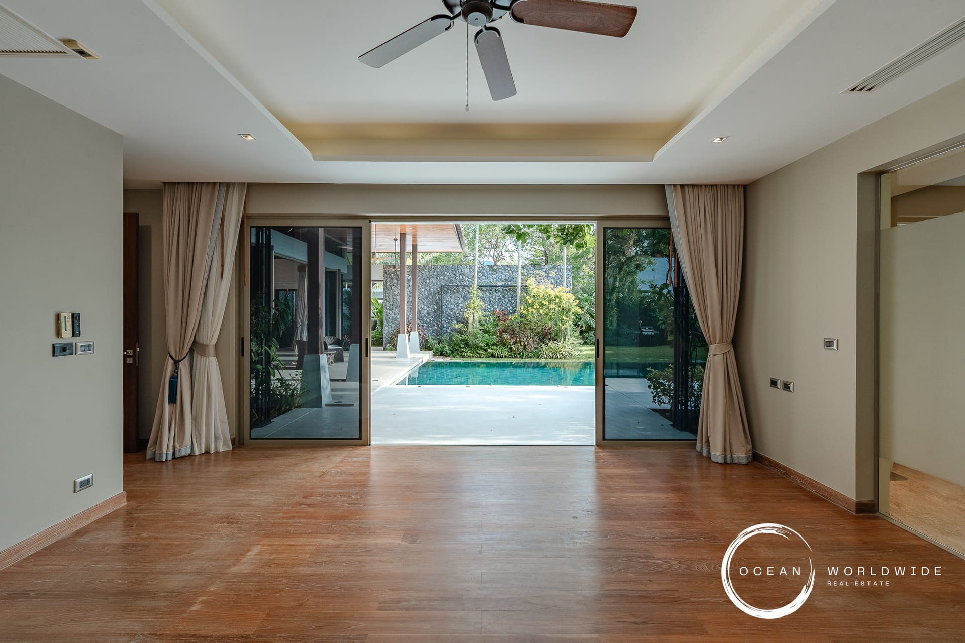 Living area with wood floors and large sliding glass doors opening to a pool and garden, beige walls, and a ceiling fan above.