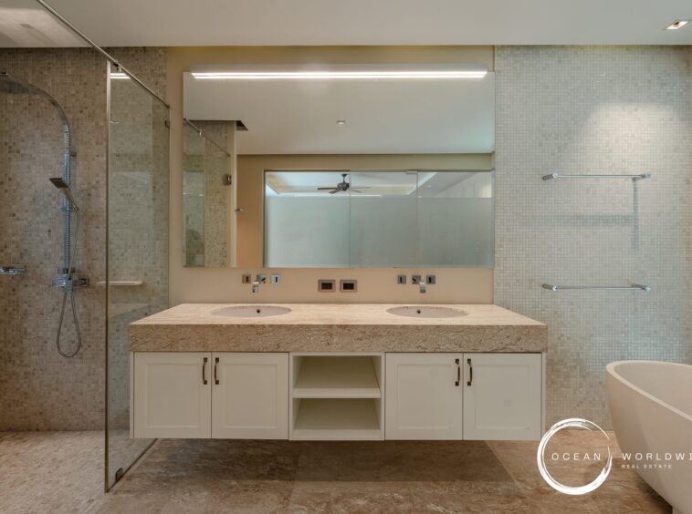 Modern bathroom with a double vanity, beige granite countertop, and two sinks reflected in a large mirror; glass shower enclosure on the left and a freestanding tub on the right.