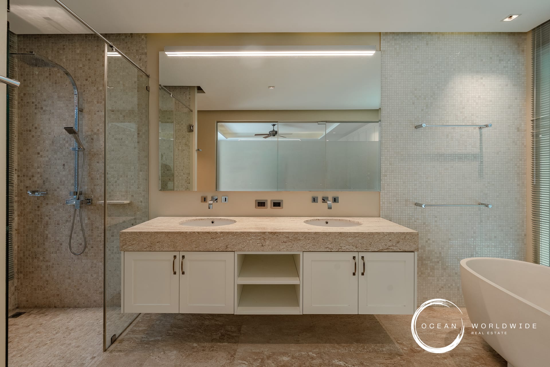 Modern bathroom with a double vanity, beige granite countertop, and two sinks reflected in a large mirror; glass shower enclosure on the left and a freestanding tub on the right.