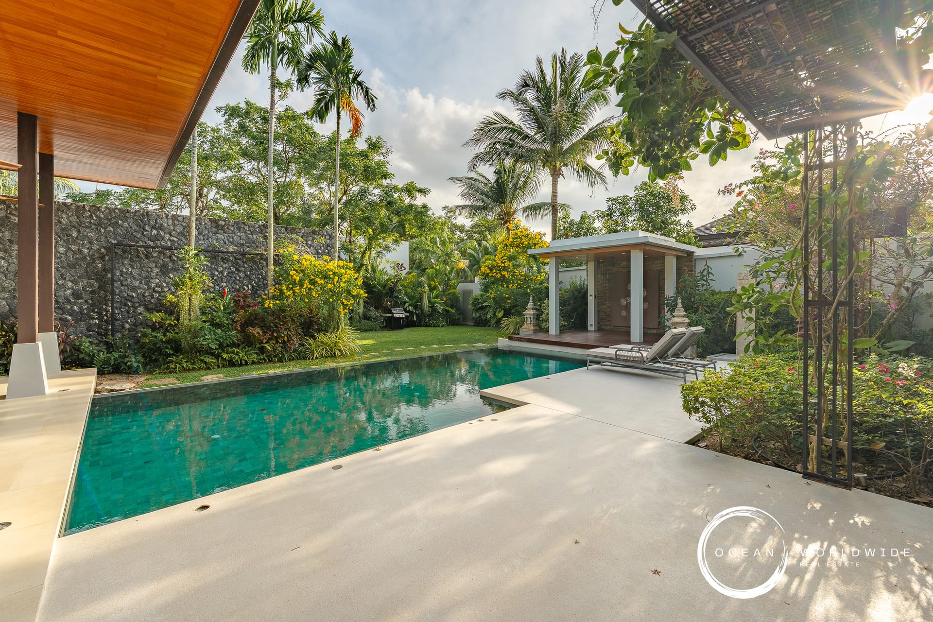 Poolside backyard with turquoise water, palm trees, and a shaded pavilion; lounge chairs on the deck nearby.