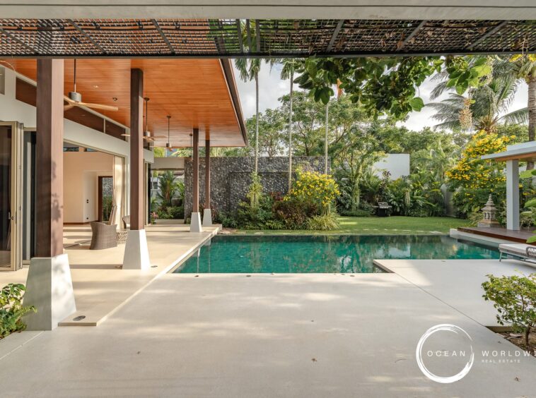 Modern outdoor living area beside a rectangular pool, with a wood-paneled ceiling, ceiling fans, and tropical plants.