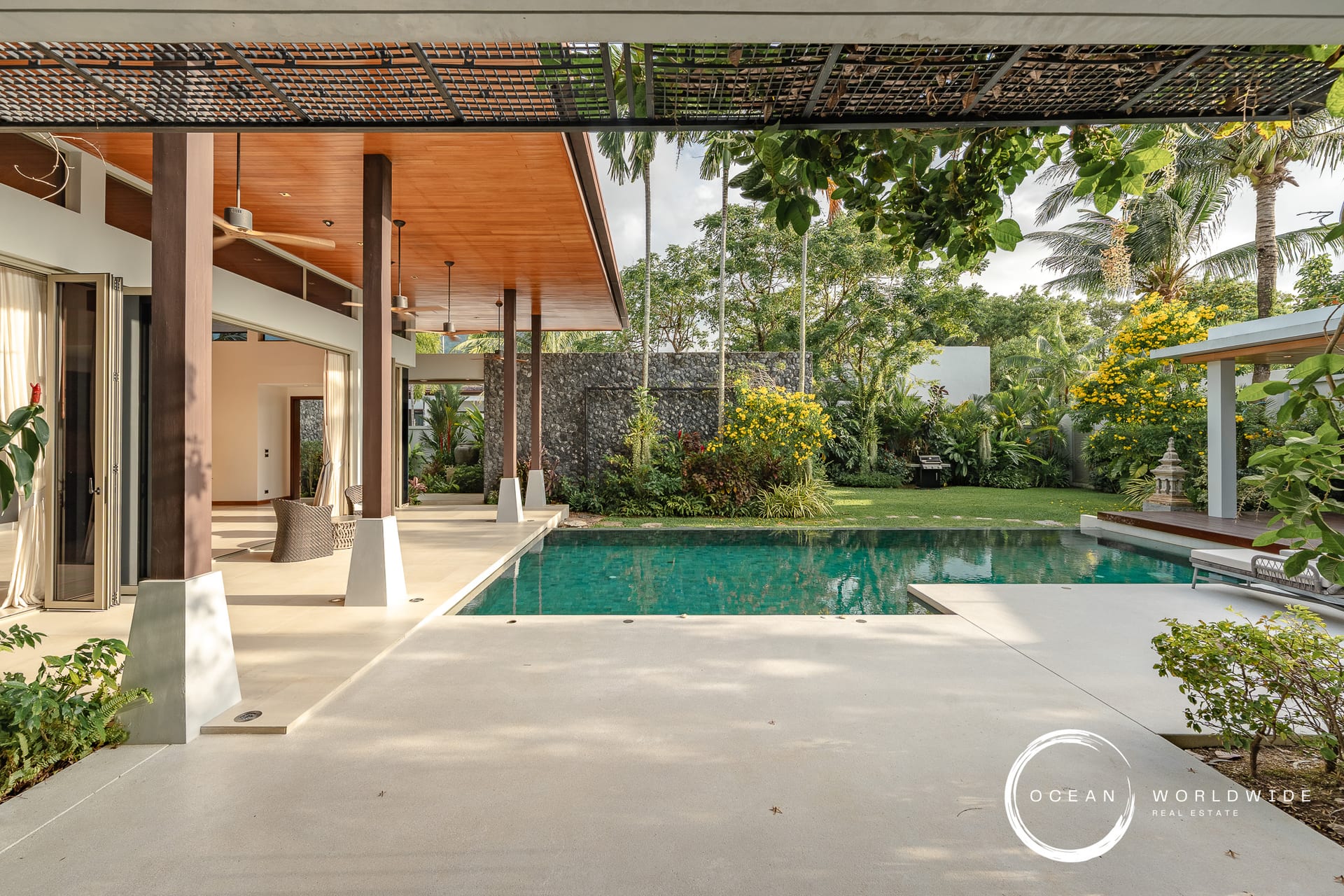 Modern outdoor living area beside a rectangular pool, with a wood-paneled ceiling, ceiling fans, and tropical plants.