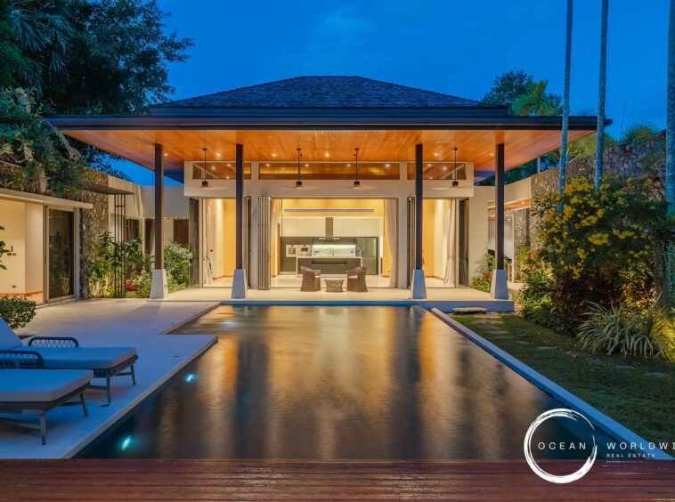 Luxurious open-air living pavilion with a lit kitchen, pool in the foreground, tropical plants at dusk, Ocean Worldwide branding.