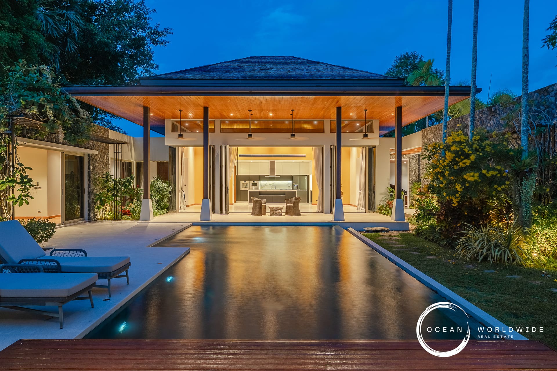 Luxurious open-air living pavilion with a lit kitchen, pool in the foreground, tropical plants at dusk, Ocean Worldwide branding.
