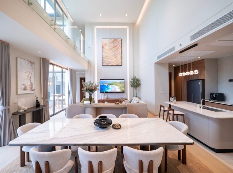 Open-plan living and dining area with a marble-topped table, beige chairs, and a kitchen island in a bright, modern home.