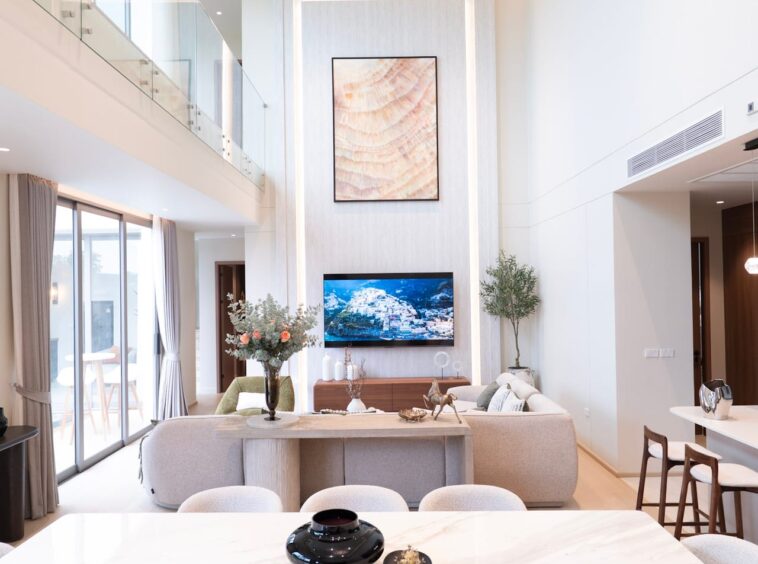 Bright open living room with a marble dining table in the foreground and beige chairs; wall-mounted TV and framed abstract artwork on a tall recessed panel in the center.